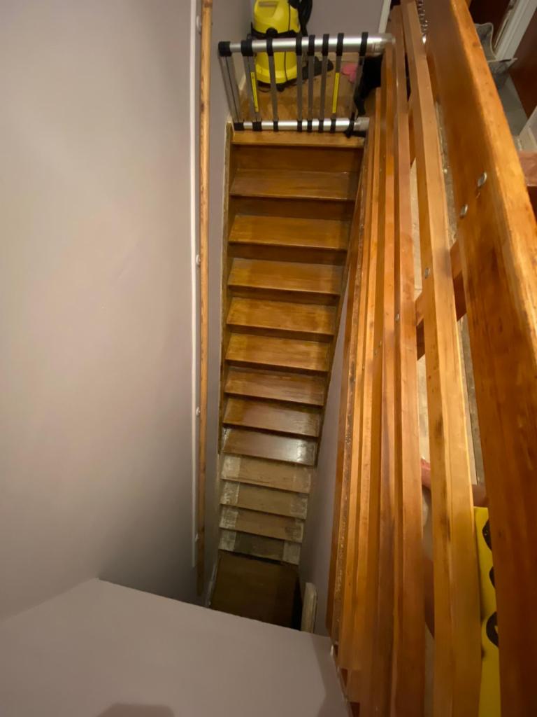 Widok z góry na drewniane schody z barierką, zabezpieczone bramką dla dziecka, z odkurzaczem w tle na górnym podeście