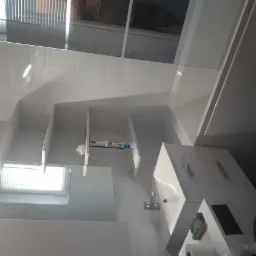 Nowoczesna, biała łazienka po remoncie. Widoczne białe płytki na ścianach, zabudowana wanna z parawanem, szafka z umywalką, lustro i półki z akcesoriami łazienkowymi.