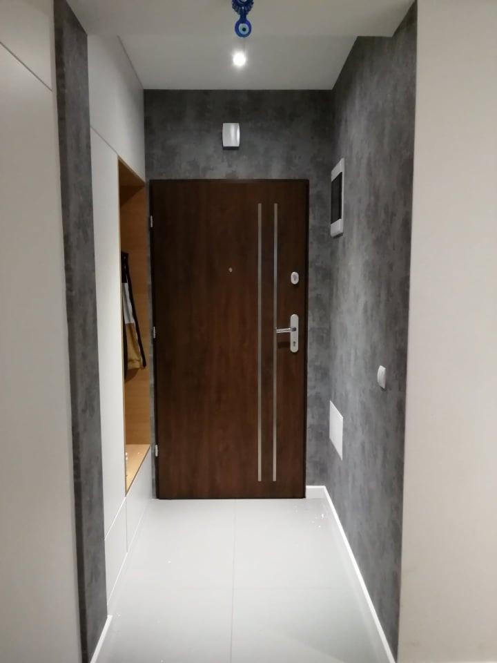 Wnętrze przedpokoju po remoncie: brązowe drzwi wejściowe z dwoma srebrnymi paskami na tle ściany z efektem betonu, białe płytki podłogowe, zabudowa szafy z wnęką i wieszakami, dekoracyjny amulet...