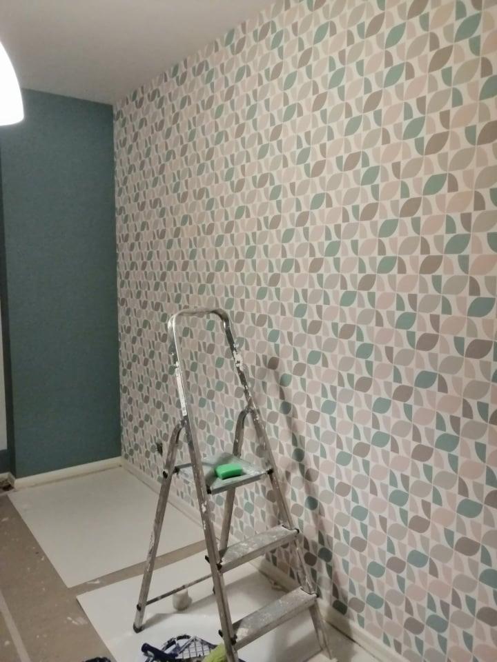 Ściana pokryta tapetą w geometryczny wzór w odcieniach beżu, szarości i zieleni; drabina aluminiowa z zieloną gąbką na jednym ze stopni, częściowo widoczna niebieska ściana po lewej stronie.