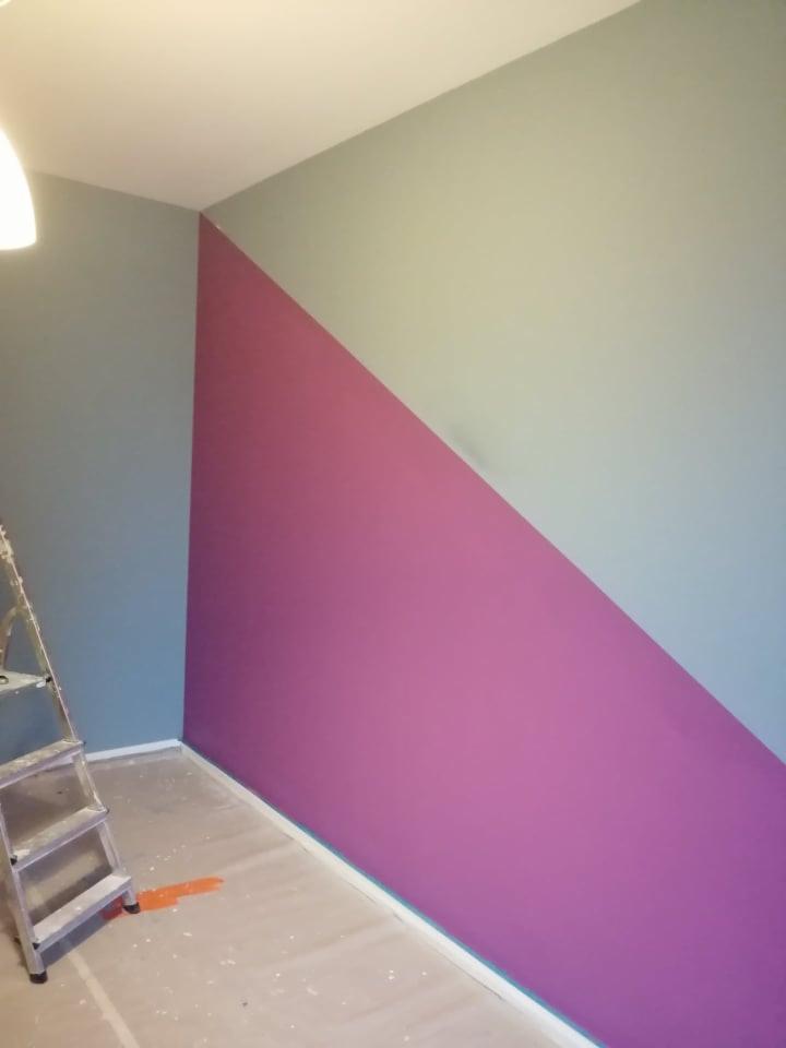 Ściana pomalowana w geometryczny wzór z użyciem fioletu i szaro-zielonego, widoczna drabina malarska i plama farby na podłodze.