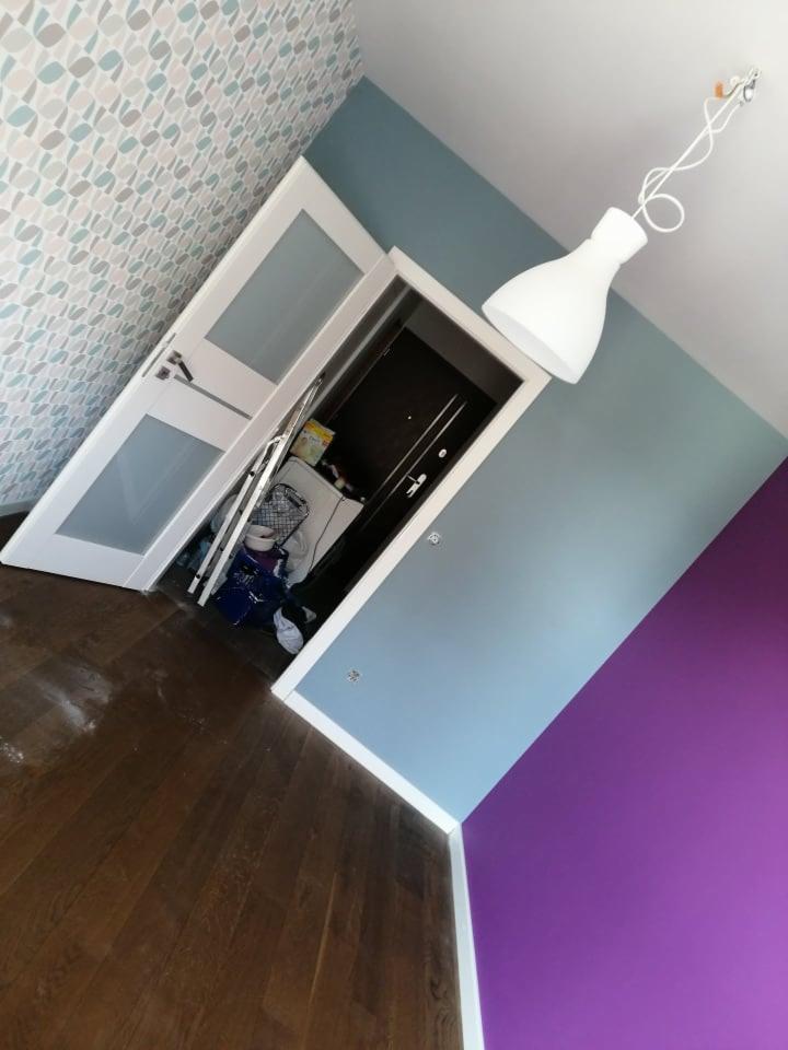 Pokój po remoncie: brązowa podłoga z ciemnego drewna, ściany pomalowane na niebiesko i fioletowo, wzorzysta tapeta, białe drzwi z przeszkleniami, widoczny bałagan w przejściu, biała lampa wisząca.