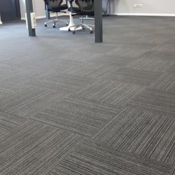 Carpet Tomasz Kurkowski - Biuro z szarą wykładziną dywanową w kwadraty, widoczne biurka i krzesła w tle.