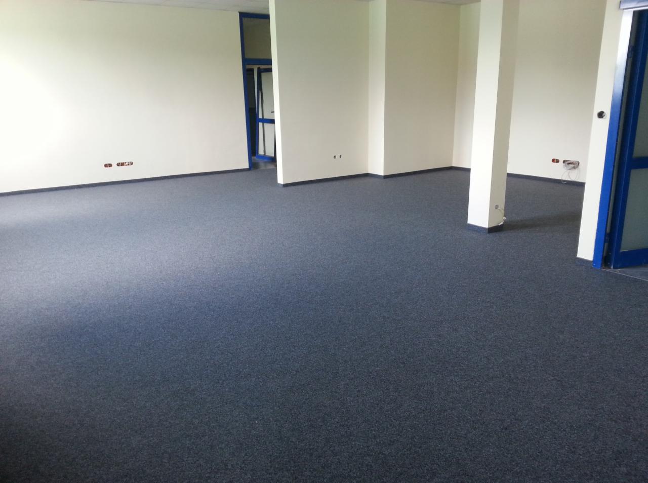 Nowa, ciemnoszara wykładzina dywanowa w pustym biurze z białymi ścianami i kolumnami, widoczne gniazdka elektryczne i fragmenty niebieskich drzwi.