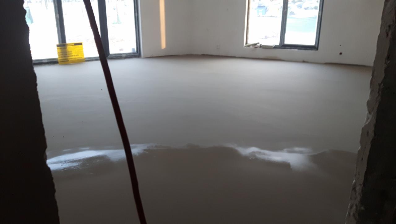 Wylewka samopoziomująca w jasnym pomieszczeniu z dwoma oknami, widoczne ślady wilgoci i żółta tablica informacyjna w tle.