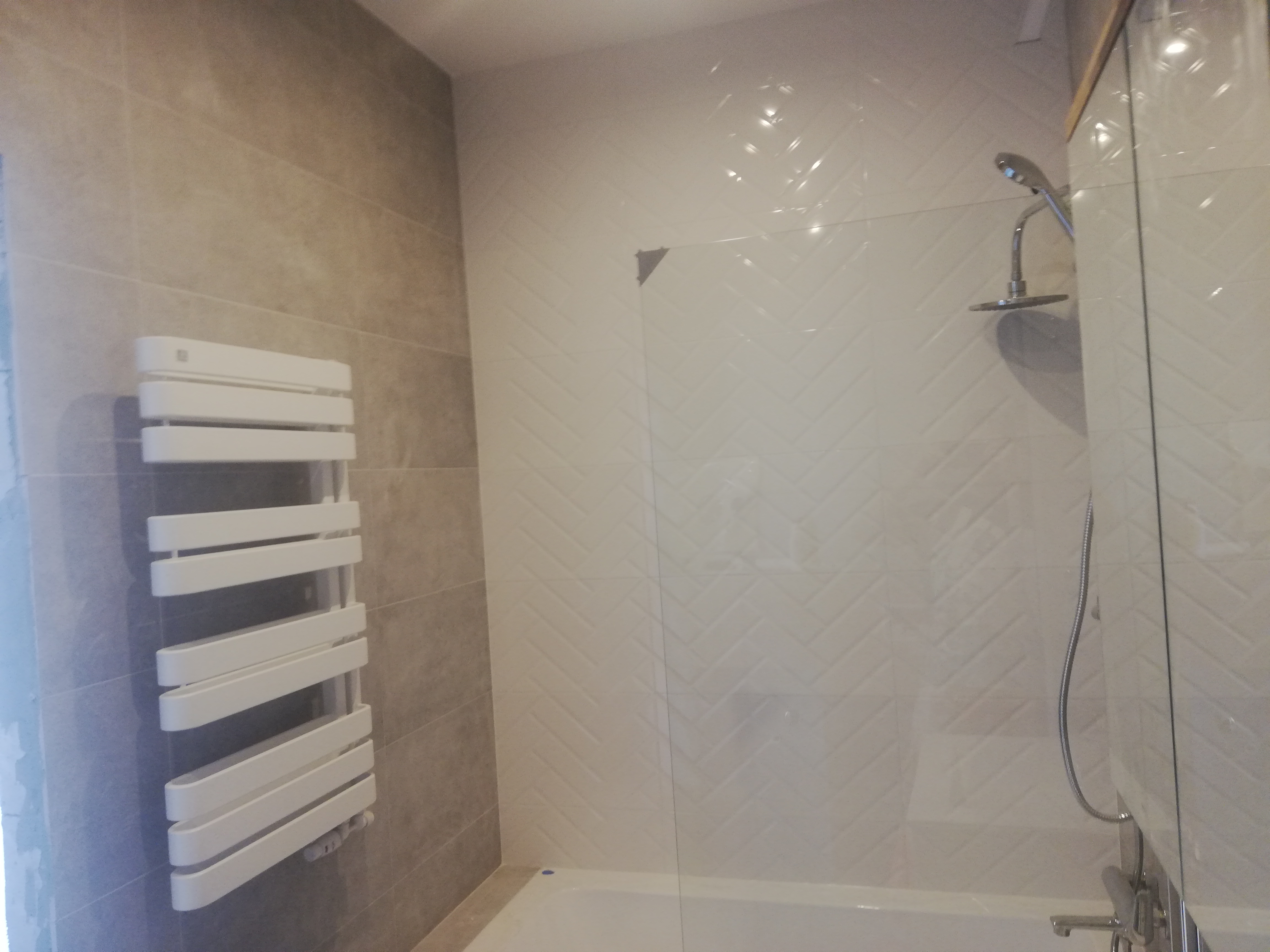 Nowoczesna łazienka z grzejnikiem drabinkowym na ścianie wyłożonej beżowymi płytkami oraz kabiną prysznicową z transparentną szybą i białymi płytkami ułożonymi w jodełkę.