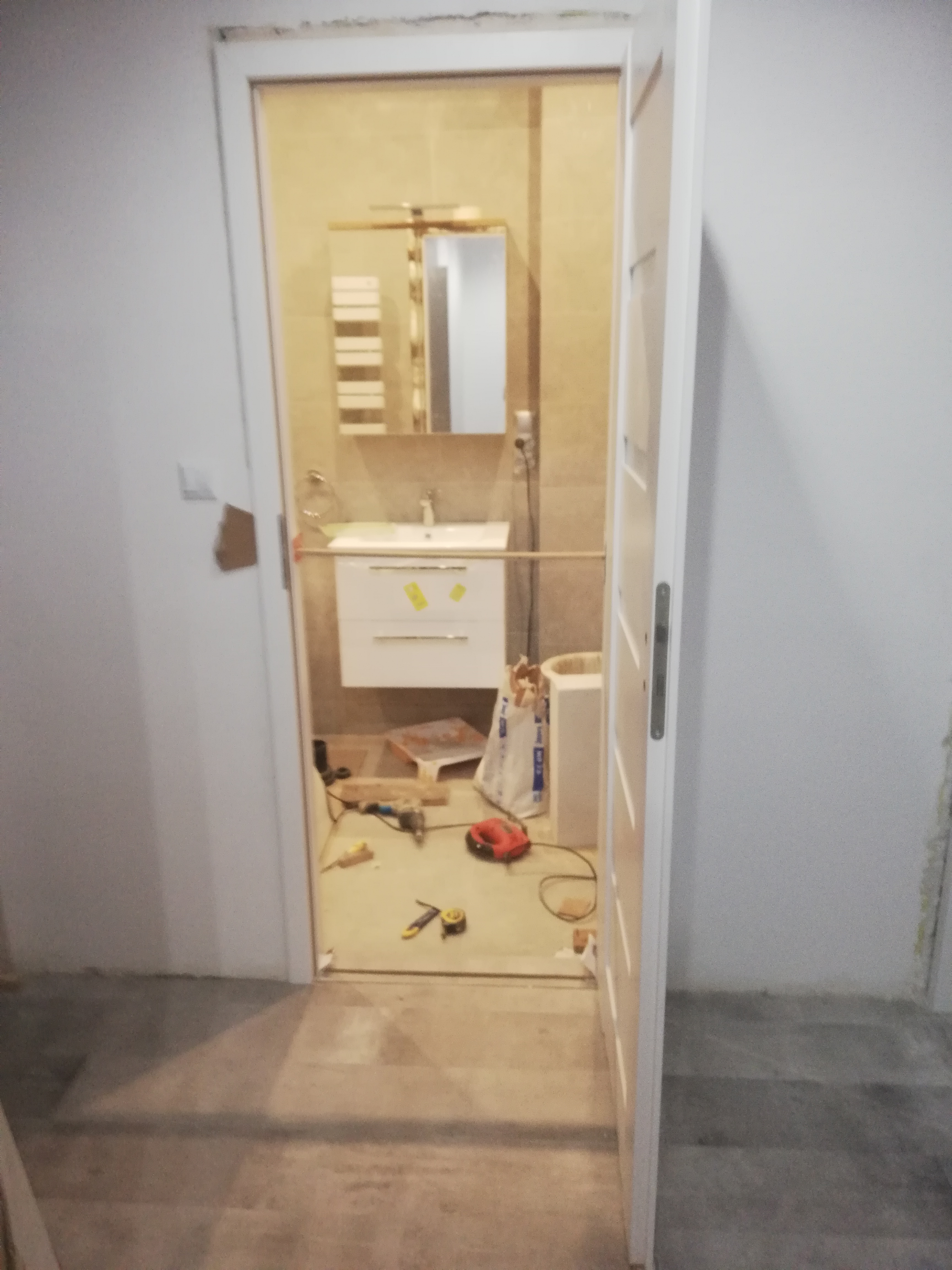 Remont łazienki w trakcie: widok przez otwarte drzwi na zamontowaną umywalkę z szafką, lustro z oświetleniem i narzędzia na podłodze.