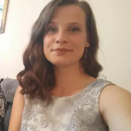 Portret uśmiechniętej kobiety z brązowymi włosami i delikatnym makijażem, ubrana w sukienkę z koronkowymi aplikacjami i kolczyki.