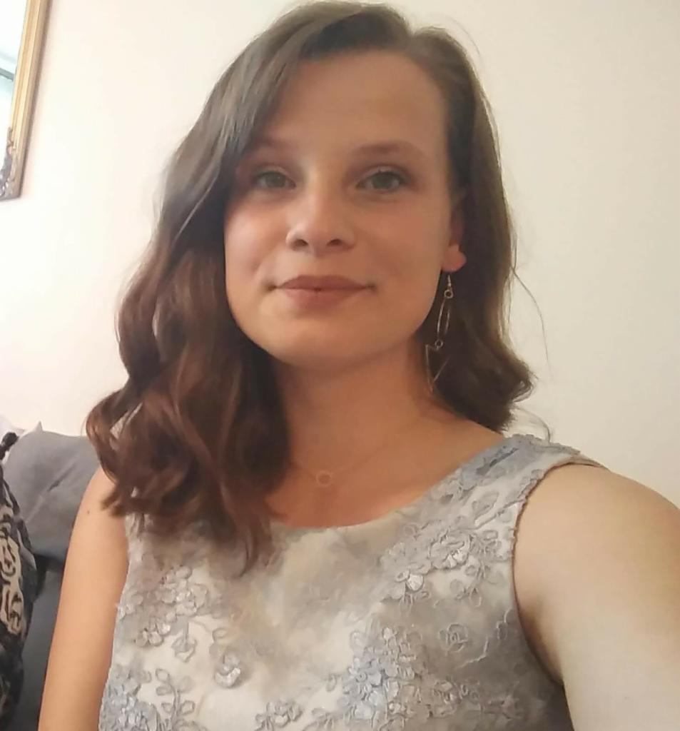 Portret uśmiechniętej kobiety z brązowymi włosami i delikatnym makijażem, ubrana w sukienkę z koronkowymi aplikacjami i kolczyki.