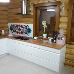 Nowoczesna kuchnia w drewnianym domu z białymi szafkami, drewnianym blatem i panelem dekoracyjnym nad płytą grzewczą z motywami ludowymi.