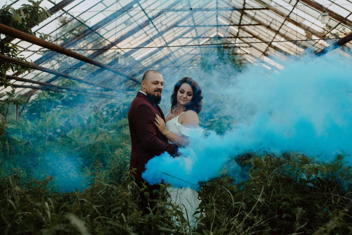 Para młoda w objęciach w szklarni, otoczona niebieskim dymem, on w bordowym garniturze, ona w białej sukni ślubnej.