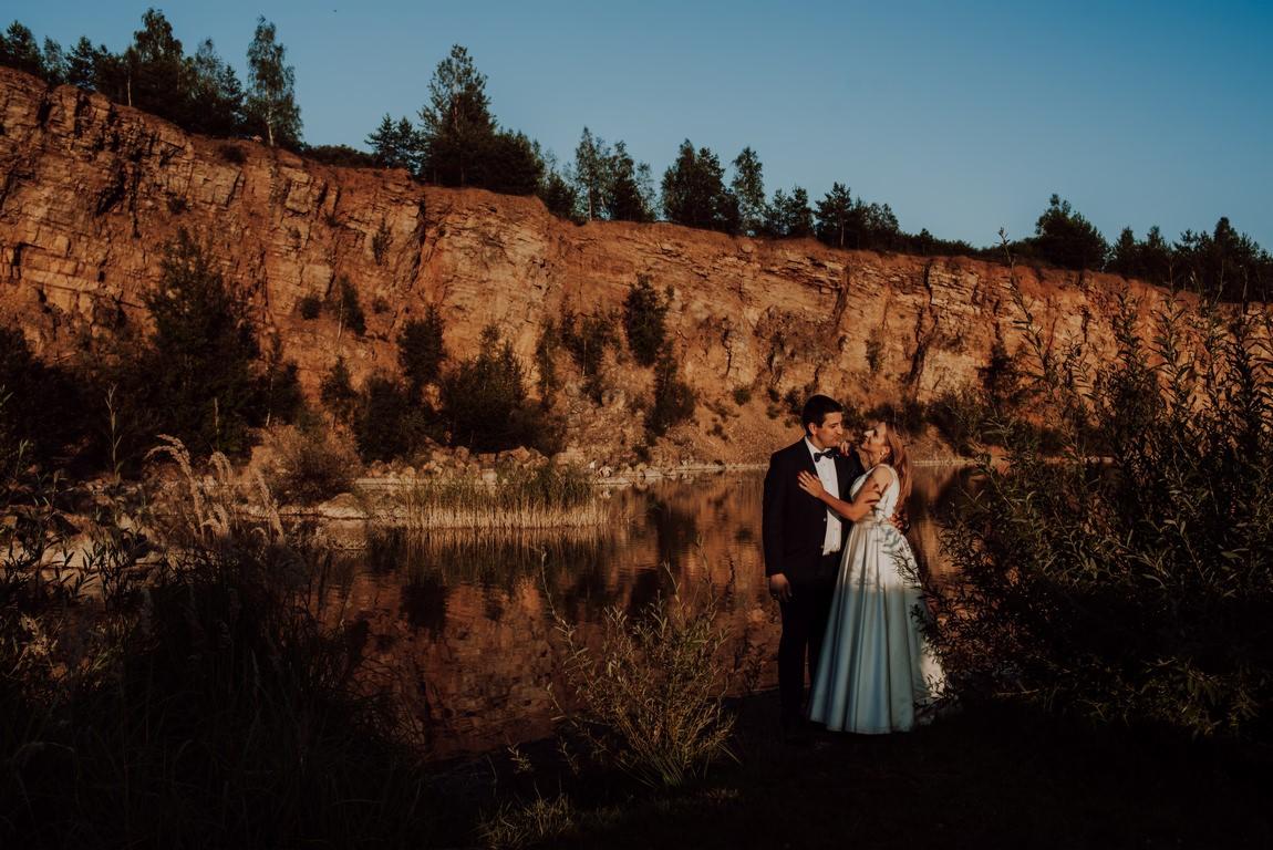 Para młoda w eleganckich strojach, obejmująca się czule na tle malowniczego krajobrazu z jeziorem i skalistym klifem, oświetlonych zachodzącym słońcem.