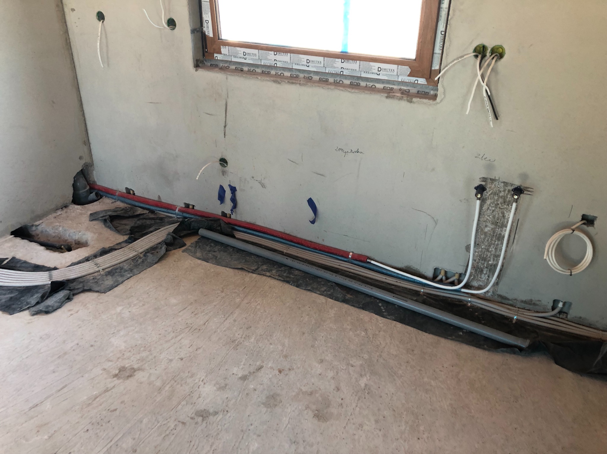 Surowy stan wnętrza z widocznymi instalacjami hydraulicznymi i elektrycznymi w trakcie montażu, ściany w stanie szpachli, okno z naklejkami producenta.