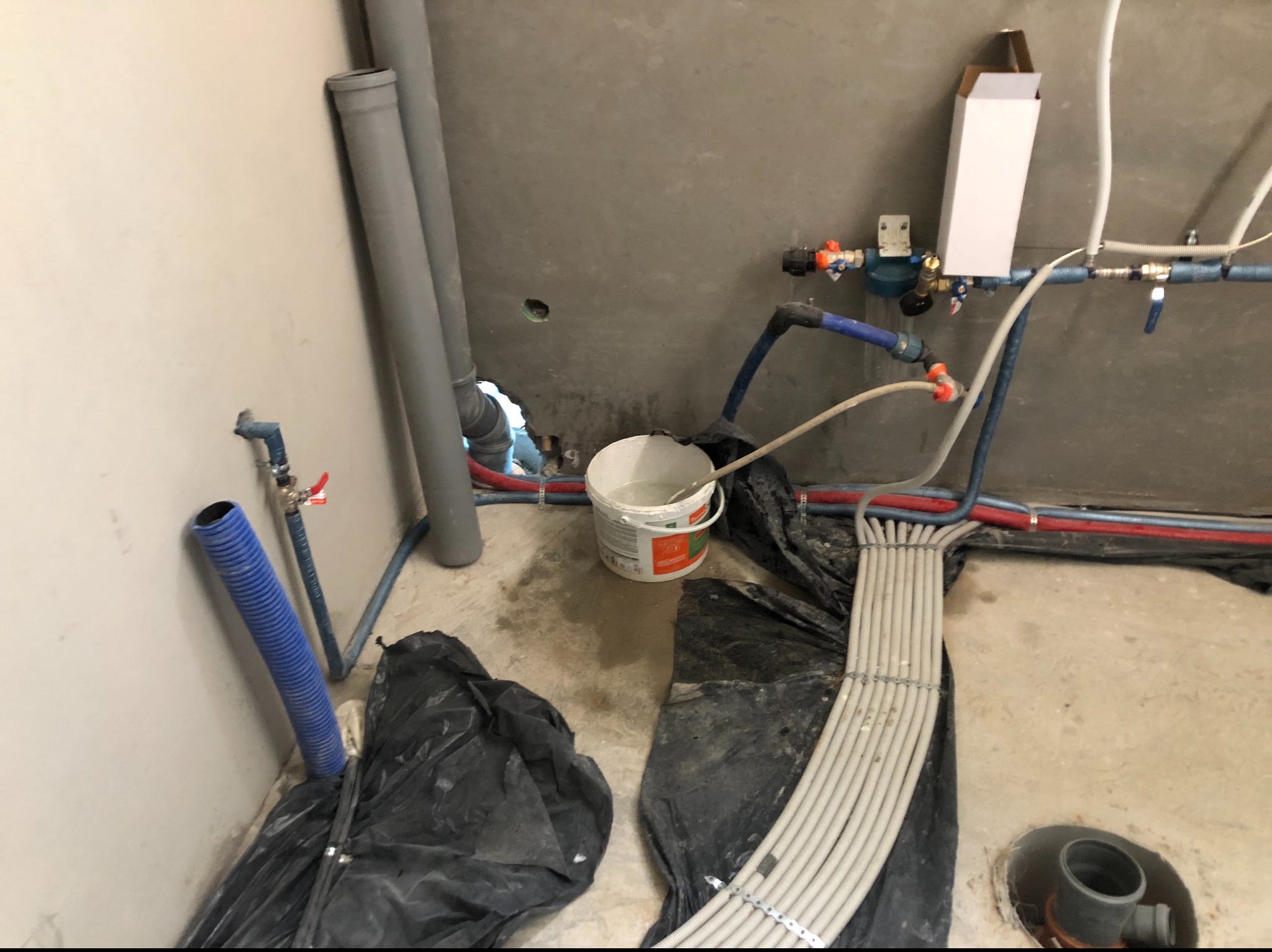 Surowy stan wykończenia wnętrza z widocznymi rurami instalacji wodnej i kanalizacyjnej, zabezpieczonymi folią elementami oraz wiadrem z zawartością, prawdopodobnie zaprawą.