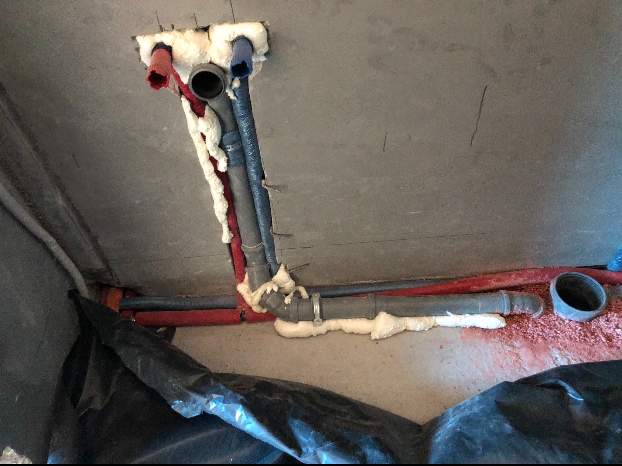 Widok na instalację rur w stropie betonowym, zabezpieczonych pianką montażową, z widocznymi rurami w kolorach czerwonym i niebieskim oraz czarną folią ochronną na podłodze.