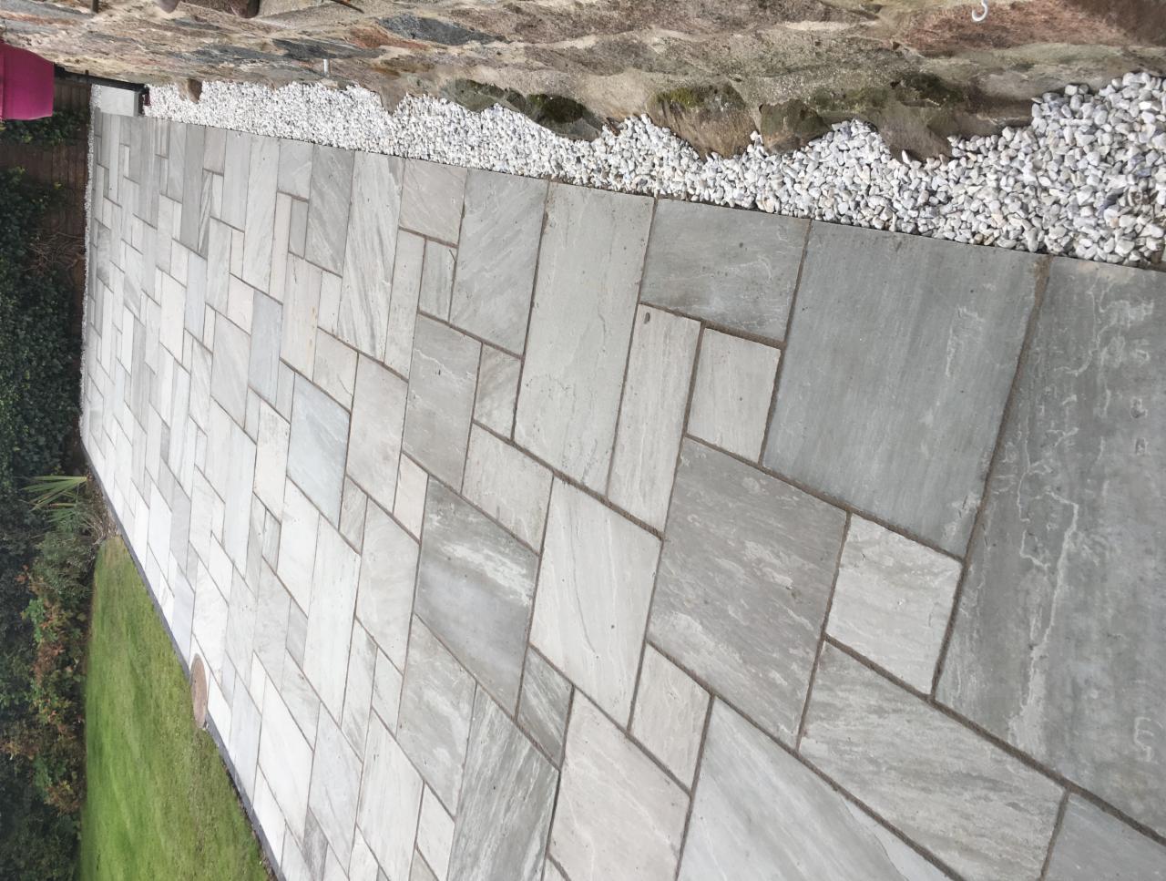 Ścieżka z jasnoszarych, prostokątnych płyt ułożonych w nieregularny wzór, obok ściany z kamienia i białych kamyków.