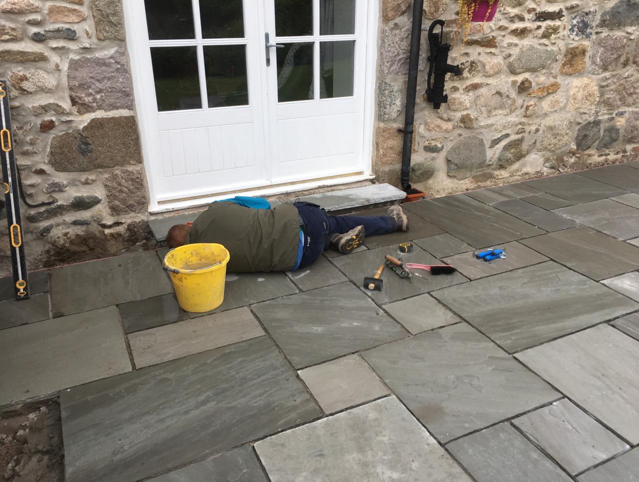 Pracownik w kurtce leży na ułożonej, szarej kostce brukowej przed białymi drzwiami, obok żółtego wiadra i narzędzi.