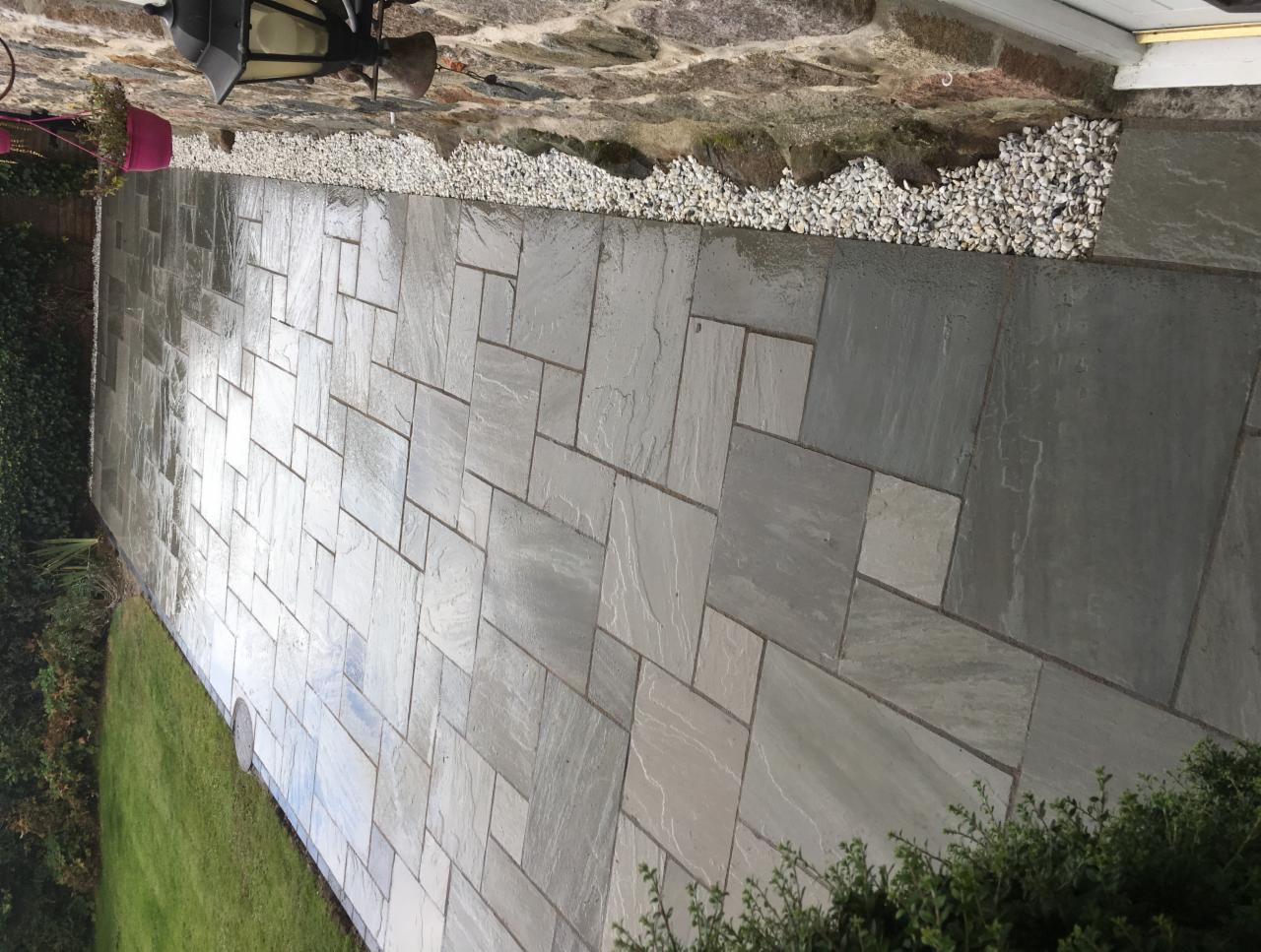 Świeżo ułożona, wilgotna ścieżka z szarej kostki, obramowana białym żwirem i zielonym trawnikiem, prowadząca do wejścia z oświetleniem.