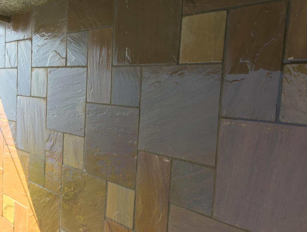 Świeżo ułożona, mokra nawierzchnia z nieregularnych, prostokątnych płyt kamiennych w odcieniach brązu i szarości, z widocznym odbiciem światła słonecznego.