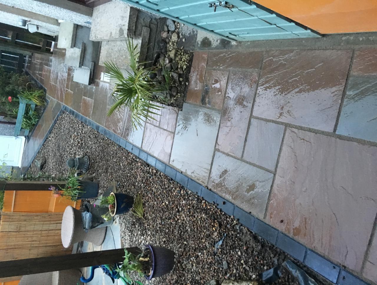 Świeżo ułożony chodnik z nieregularnych, brązowo-szarych płyt kamiennych, oddzielony od rabaty z drobnym żwirem czarną obwódką. Na rabacie rosną rośliny, w tym mała palma. Widoczne schody wejściowe.