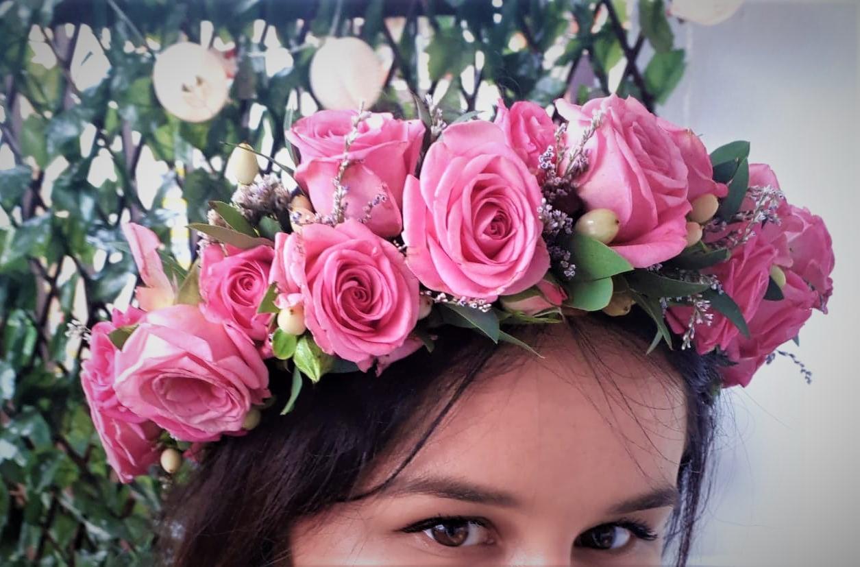 Kobieta z bliska, w wianku z różowych róż, z zielonymi liśćmi i małymi białymi jagodami, na tle dekoracji z zielonych gałązek i światełek.