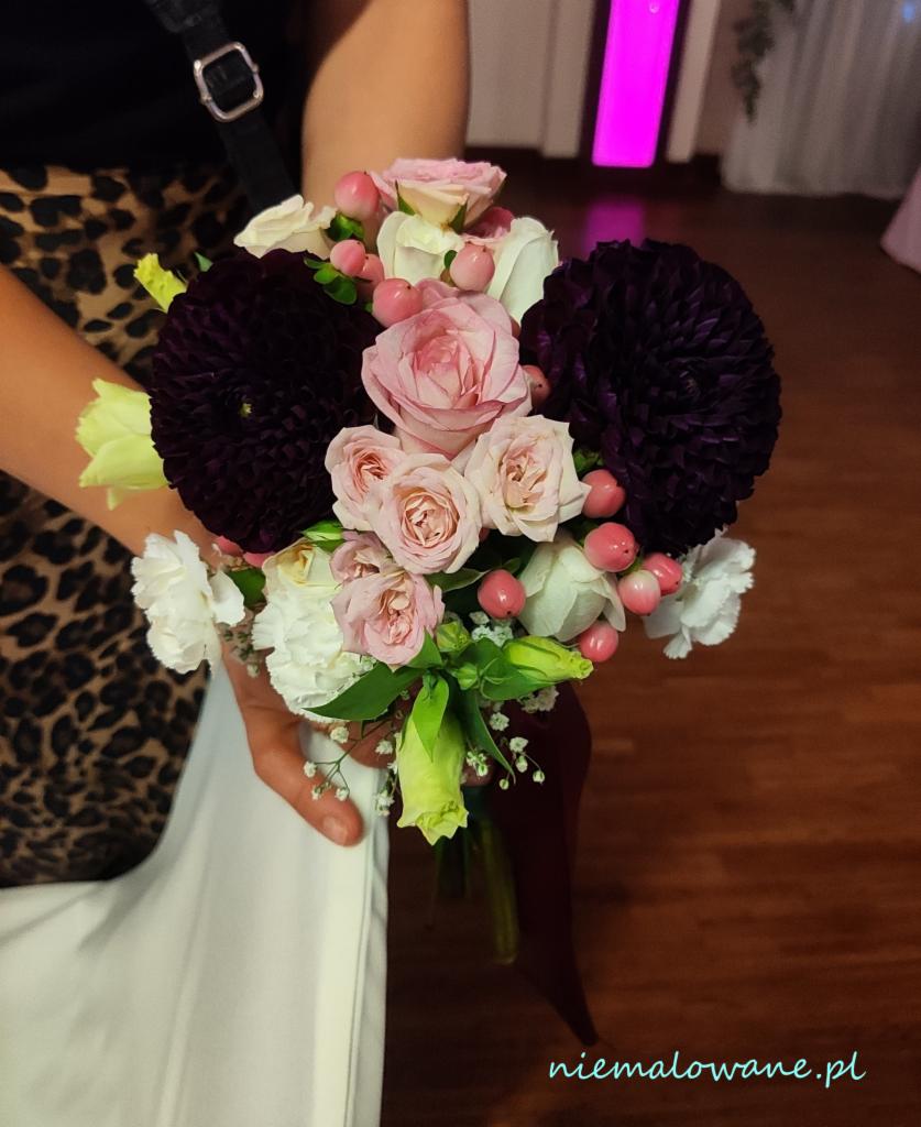 Delikatny bukiet ślubny z ciemnofioletowych dalii, różowych róż i białych goździków, trzymany w dłoniach na tle wzorzystej sukienki.