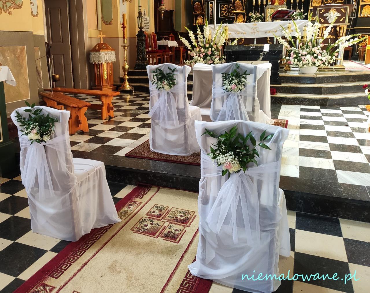 Dekoracja krzeseł w kościele na uroczystość ślubną, białe pokrowce z tiulu i kompozycje kwiatowe z róż i zielenią.