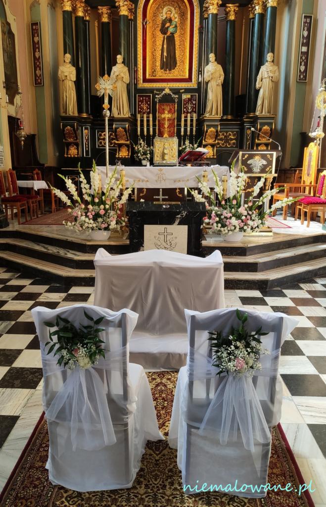 Dekoracja kościoła na ślub: białe pokrowce i kwiatowe kompozycje na krzesłach pary młodej, ołtarz z kwiatami w tle.