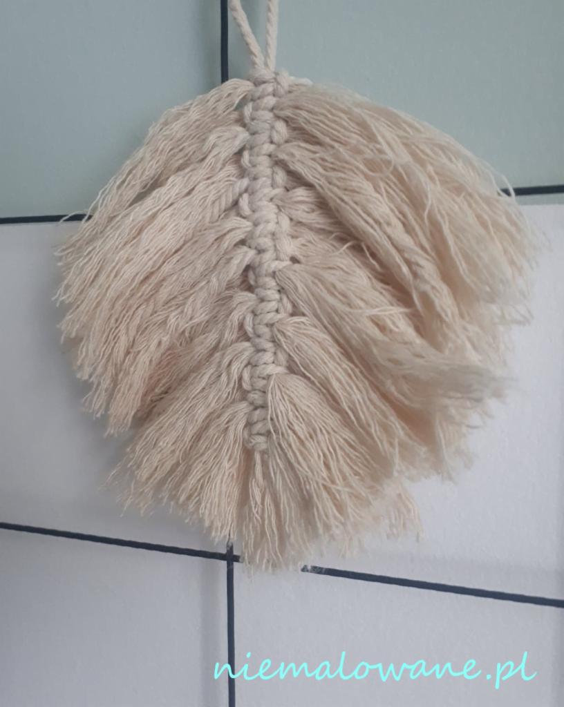 Dekoracja ścienna w formie pióra, wykonana z beżowego sznurka techniką makramy, zawieszona na ścianie z białych płytek.
