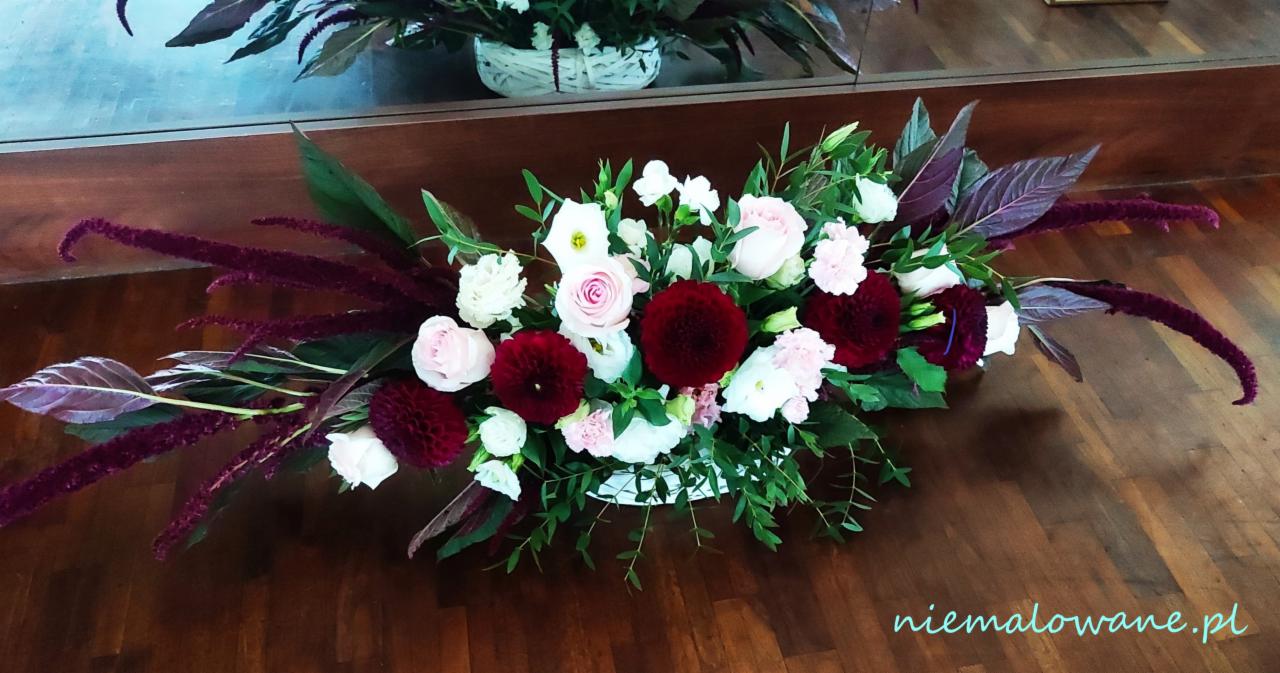 Dekoracja kwiatowa stołu z burgundowymi daliami, różowymi różami i białymi goździkami, uzupełniona amarantusem zwisającym, umieszczona w niskim, białym koszyku, na drewnianym blacie.