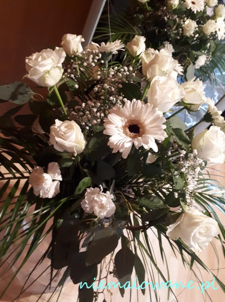 Kompozycja kwiatowa z białych róż, gerber i gipsówki z zielonymi liśćmi palmowymi, prawdopodobnie dekoracja stołu.