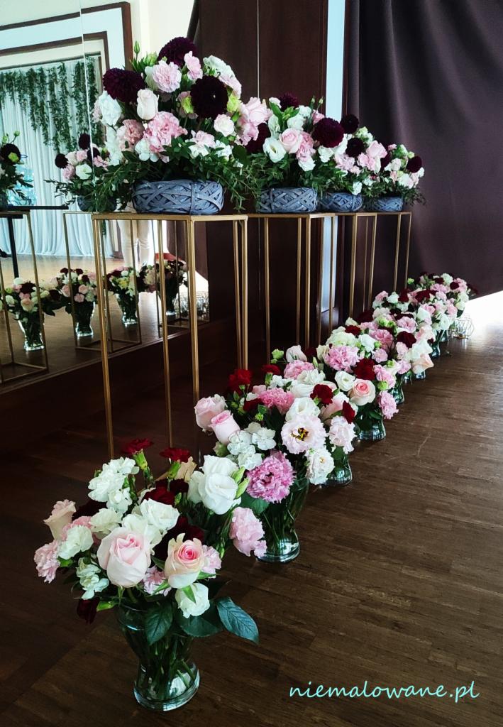 Dekoracja sali weselnej: kompozycje kwiatowe w szklanych wazonach i plecionych koszach na złotych postumentach, odbicie w lustrze, drewniana podłoga.