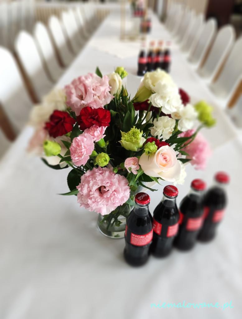 Dekoracja stołu weselnego: bukiet z różowych i białych goździków w szklanym wazonie, obok butelki Coca-Coli z czerwoną etykietą, tło z rzędem krzeseł w białych pokrowcach i długiego stołu.