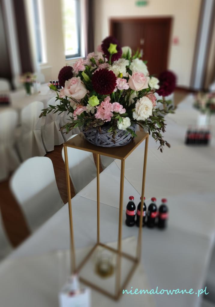 Dekoracja stołu weselnego: kompozycja kwiatowa w szarym koszyku na złotym, geometrycznym stojaku, z butelkami Coca-Coli w tle.