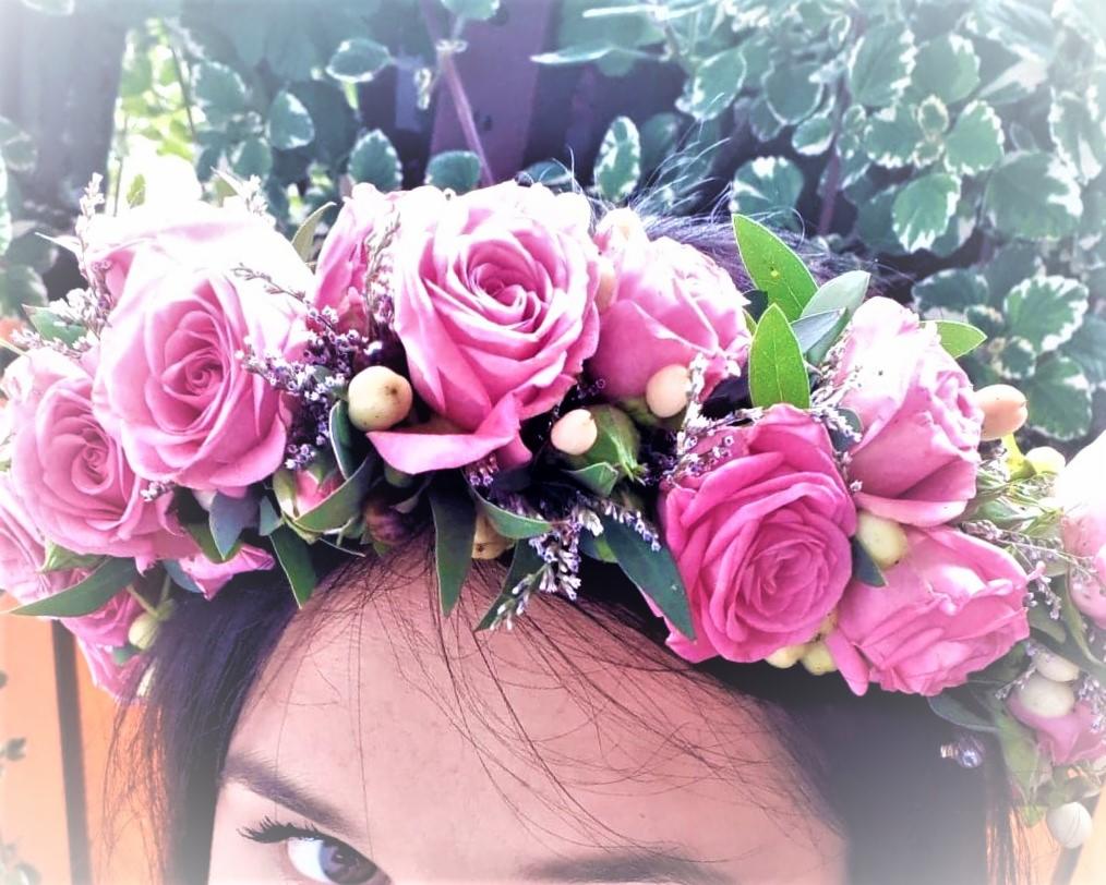 Dziewczyna z wiankiem z różowych róż i delikatnych dodatków na głowie, widoczne oczy i fragment tła z zielenią.