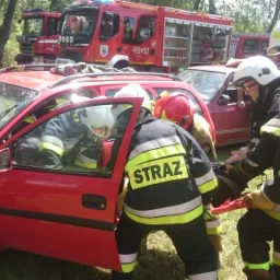 Strażacy w hełmach wyciągają poszkodowanego z rozbitego, czerwonego samochodu z popękaną szybą przednią, na tle wozów strażackich i zieleni.