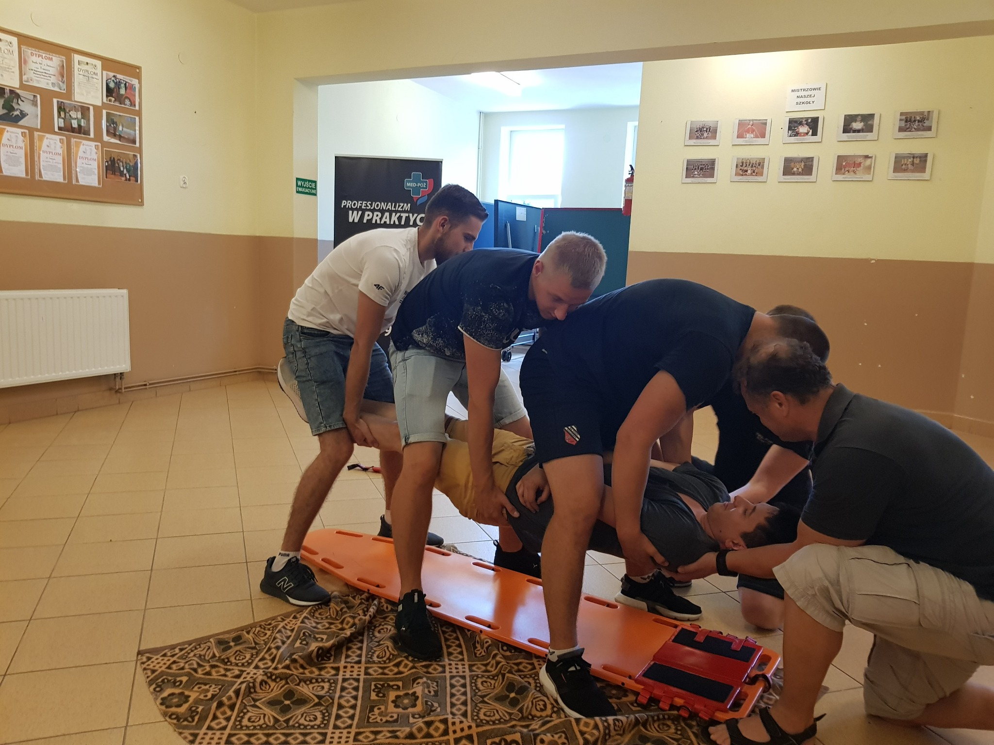 Grupa osób ćwiczy przenoszenie poszkodowanego na desce ortopedycznej w pomieszczeniu z plakatem MED-POŻ w tle.