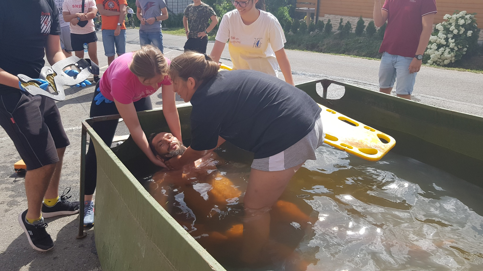 Instruktorzy demonstrują technikę ratowania topielca w zbiorniku wodnym, z użyciem deski ortopedycznej i stabilizatora głowy, podczas symulacji akcji ratowniczej na świeżym powietrzu.