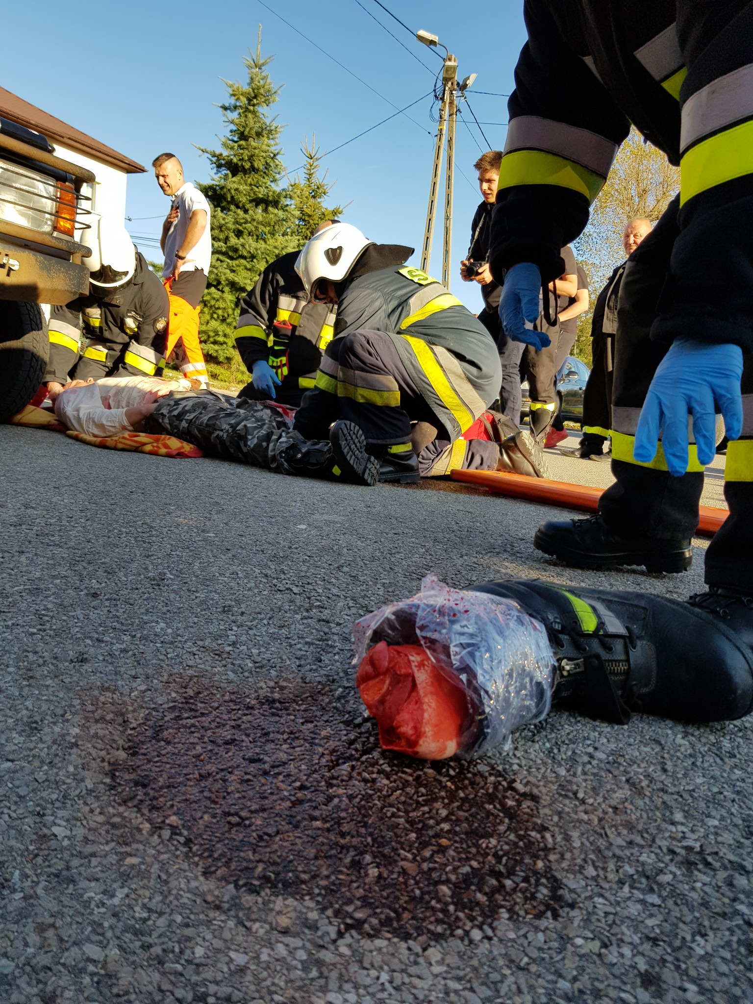 Symulacja wypadku drogowego z odciętą kończyną w worku foliowym, ratownicy medyczni udzielają pomocy poszkodowanemu leżącemu na noszach, widoczni obserwatorzy zdarzenia.
