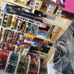 Wnętrze sklepu w Suchym Lesie z akcesoriami krawieckimi i odzieżą skórzaną, regały pełne nici, wstążek i włóczek, w pierwszym planie czarna kurtka motocyklowa.