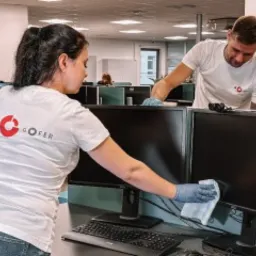 Dwoje pracowników w firmowych koszulkach GOFER czyści monitory w biurze, używając niebieskich rękawic i białej ściereczki z mikrofibry.