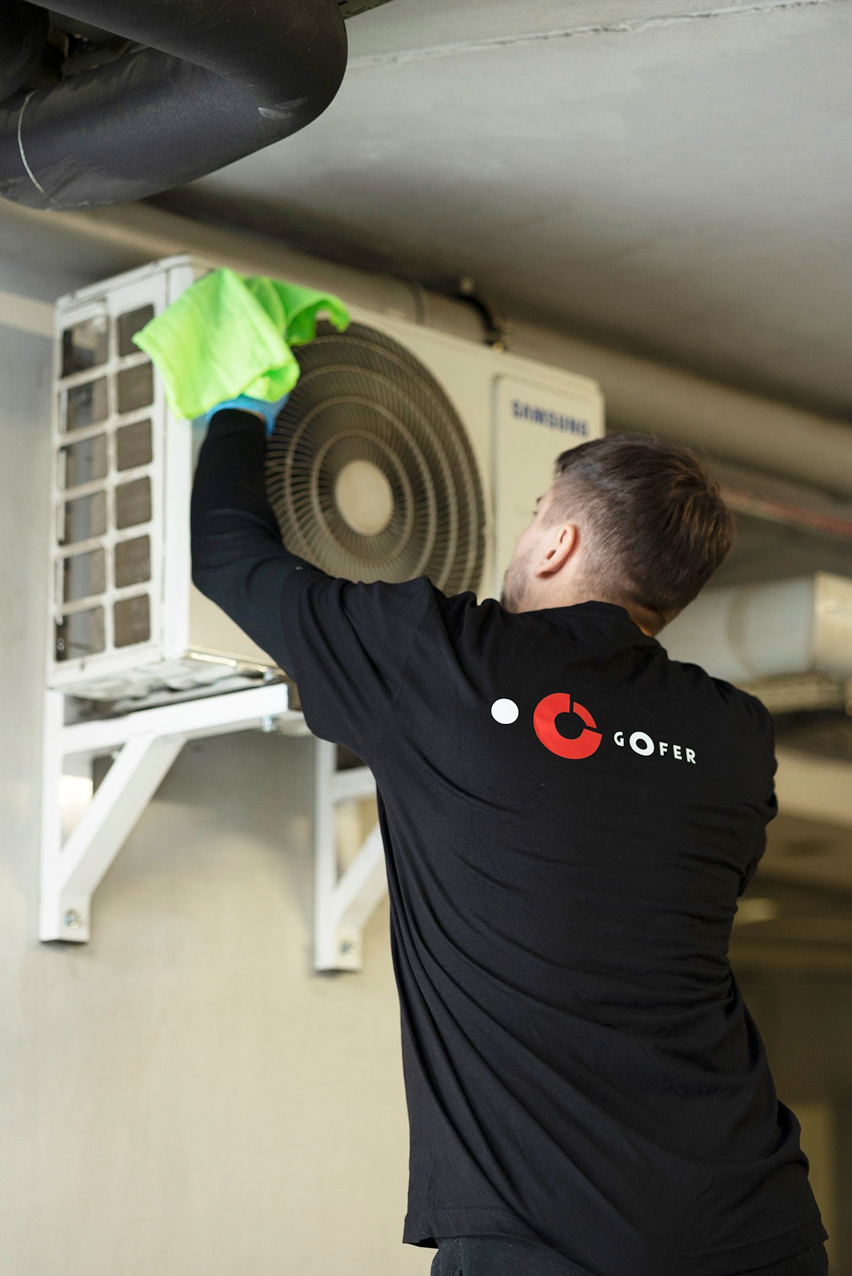 Mężczyzna w czarnej koszulce z logo firmy GOFER czyści klimatyzator Samsung, używając zielonej ściereczki i niebieskich rękawic ochronnych.