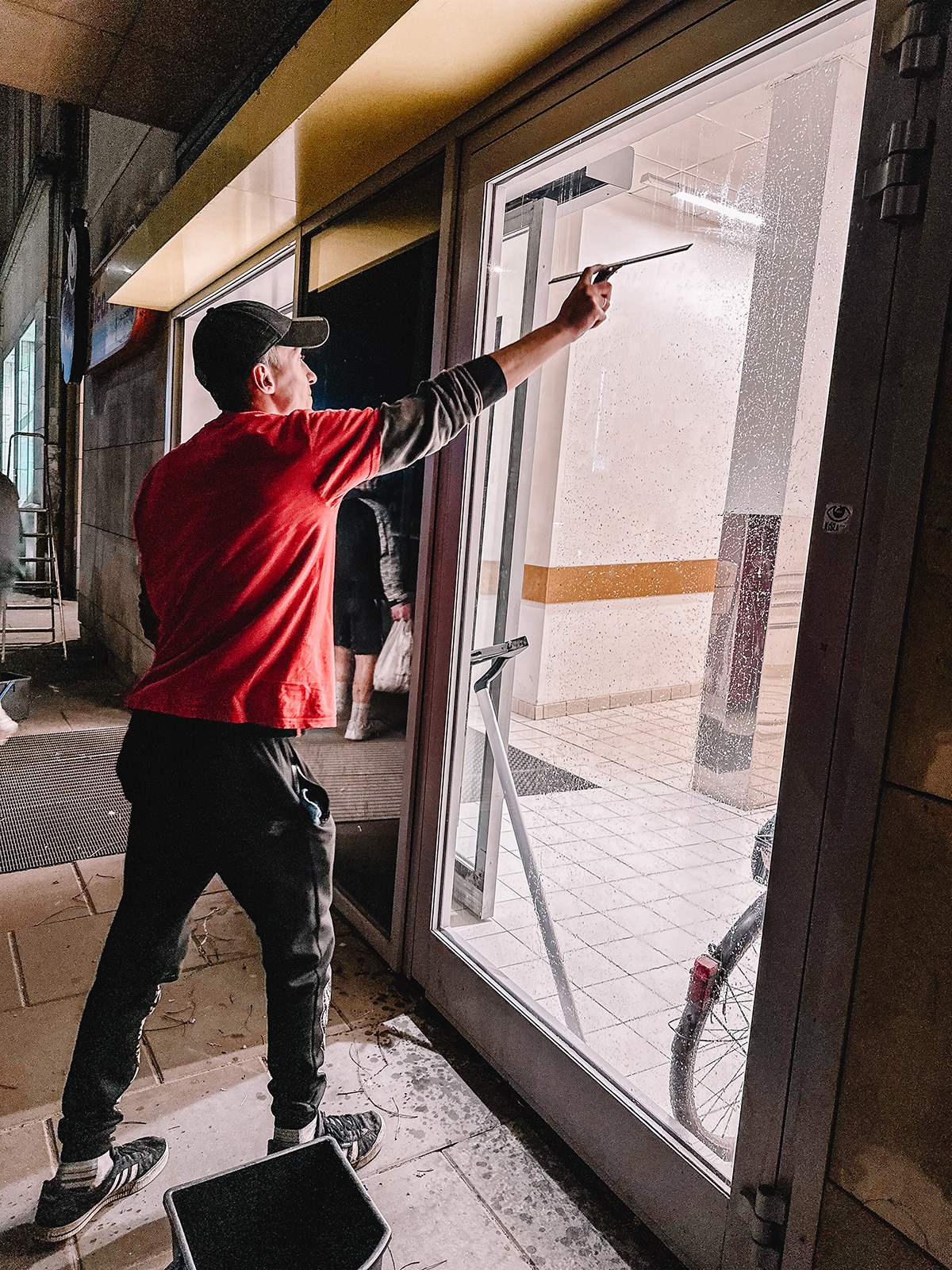 Mężczyzna w czerwonej koszulce i czapce myje szklane drzwi wejściowe do budynku za pomocą ściągaczki, widoczne krople wody na szybie, wiaderko i rower w tle.