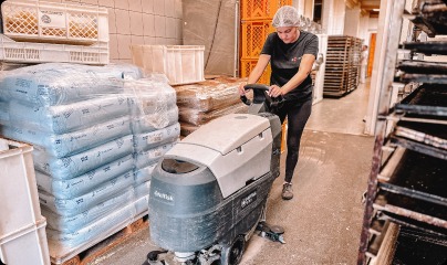 Pracownica w czepku ochronnym obsługuje automatyczną maszynę do mycia podłóg w pomieszczeniu magazynowym z paletami i regałami.