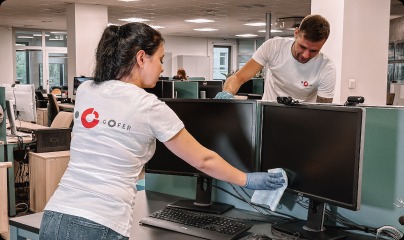 Dwoje pracowników w firmowych koszulkach GOFER czyści monitory w biurze, używając niebieskich rękawic i białej ściereczki z mikrofibry.