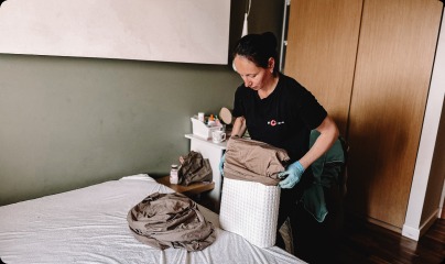 Kobieta w czarnej koszulce i niebieskich rękawiczkach układa brązową pościel na białym materacu w sypialni z zieloną ścianą i drewnianą szafą.
