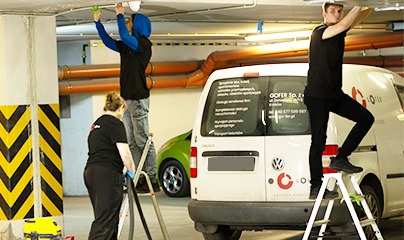 Zespół w czarnych koszulkach, stojący na drabinach i sprzątający sufit parkingu podziemnego, z widocznym samochodem dostawczym firmy.