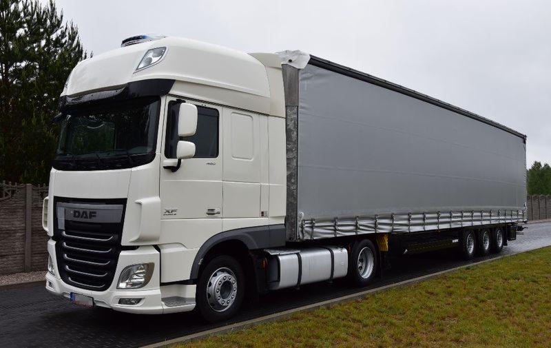 Biała ciężarówka DAF XF z szarą plandeką na naczepie, zaparkowana na poboczu z trawiastym pasem, widoczna z boku na tle pochmurnego nieba.