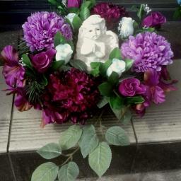 Kwiaciarnia Amarant - Kompozycja nagrobna z fioletowych i bordowych kwiatów oraz białej figurki anioła z złożonymi rękami, ułożona na płytkach.