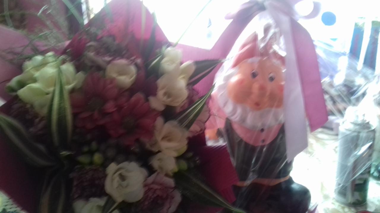 Kwiaty w różowym papierze obok zapakowanego w folię, plastikowego krasnala z białą brodą i różową czapką.